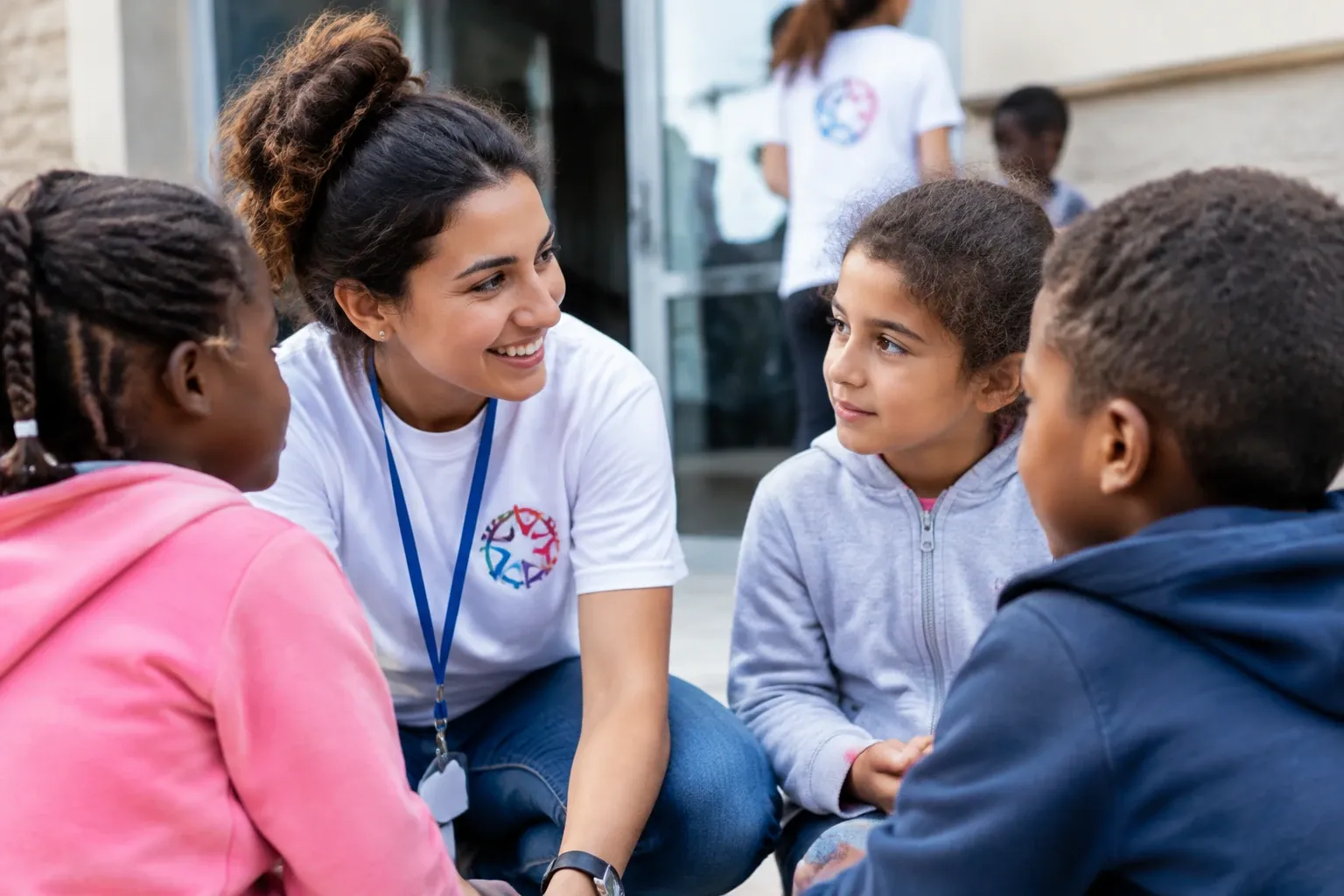 Volontaria sorridente con maglietta e badge che parla con tre bambini seduti
